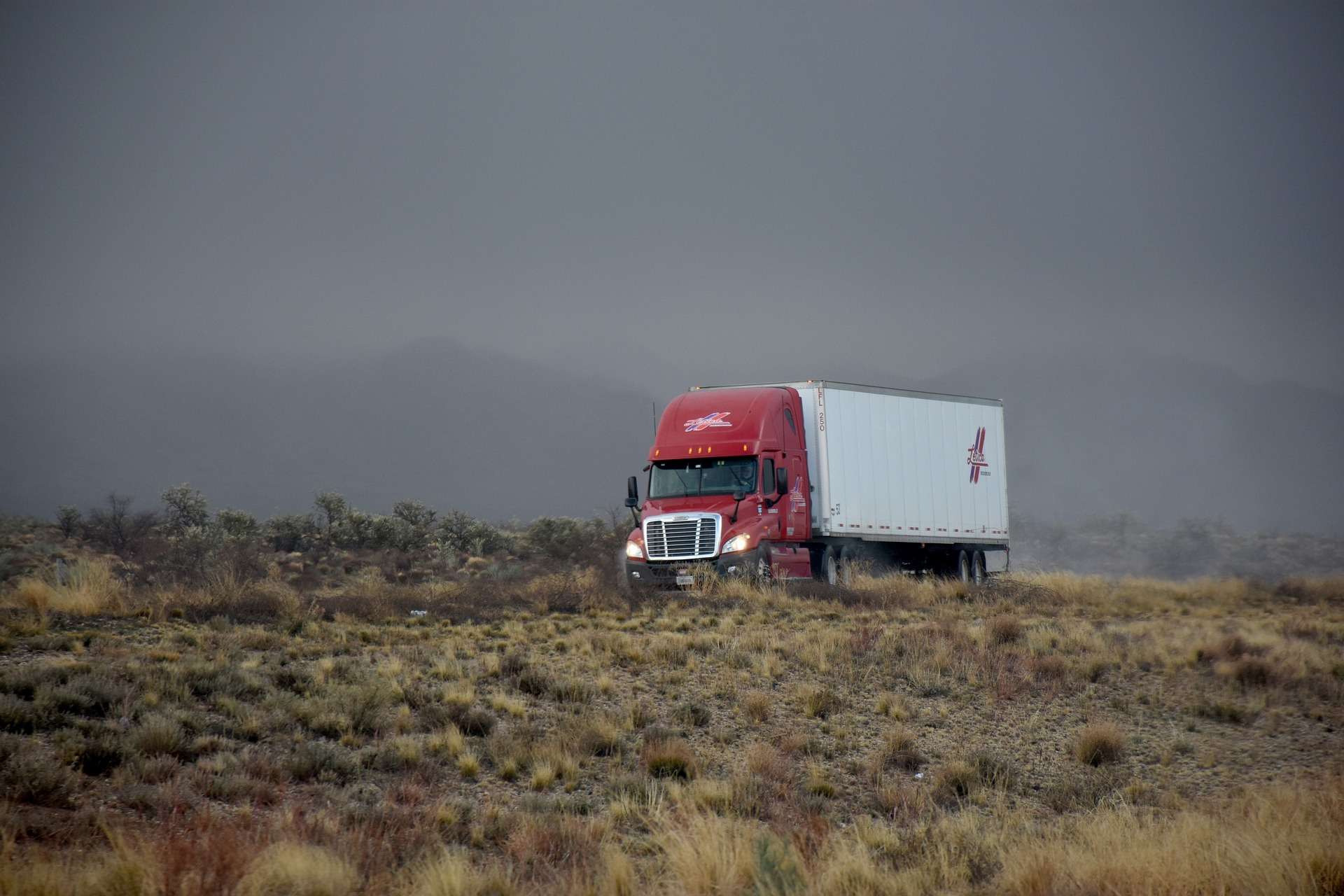 Tractocamión rojo con caja seca avanzando por un camino con cielo tormentoso
